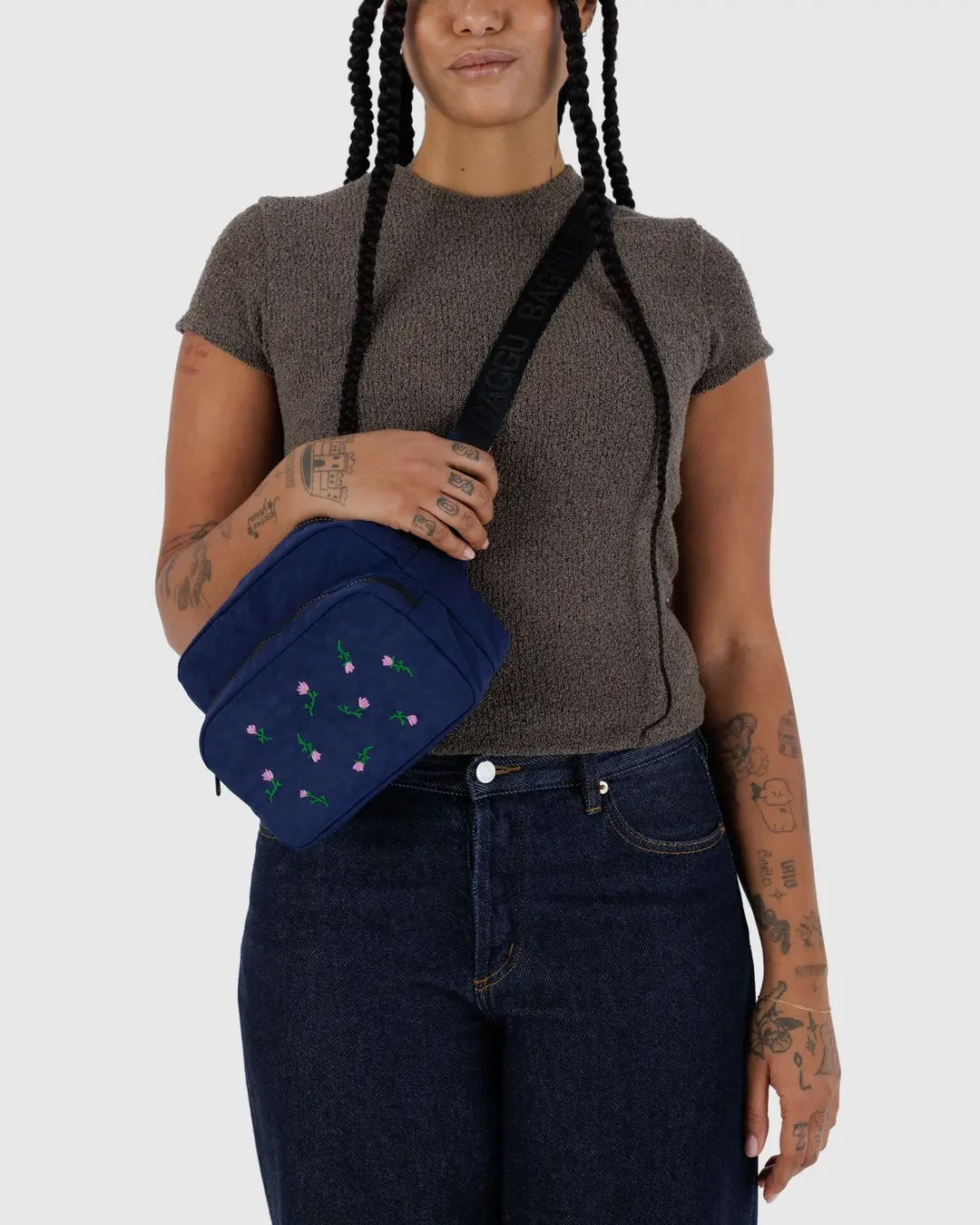 Person wearing a brown shirt and blue jeans wearing a baggu navy blue fanny pack bag with a pink embroidered floral pattern standing before a white background