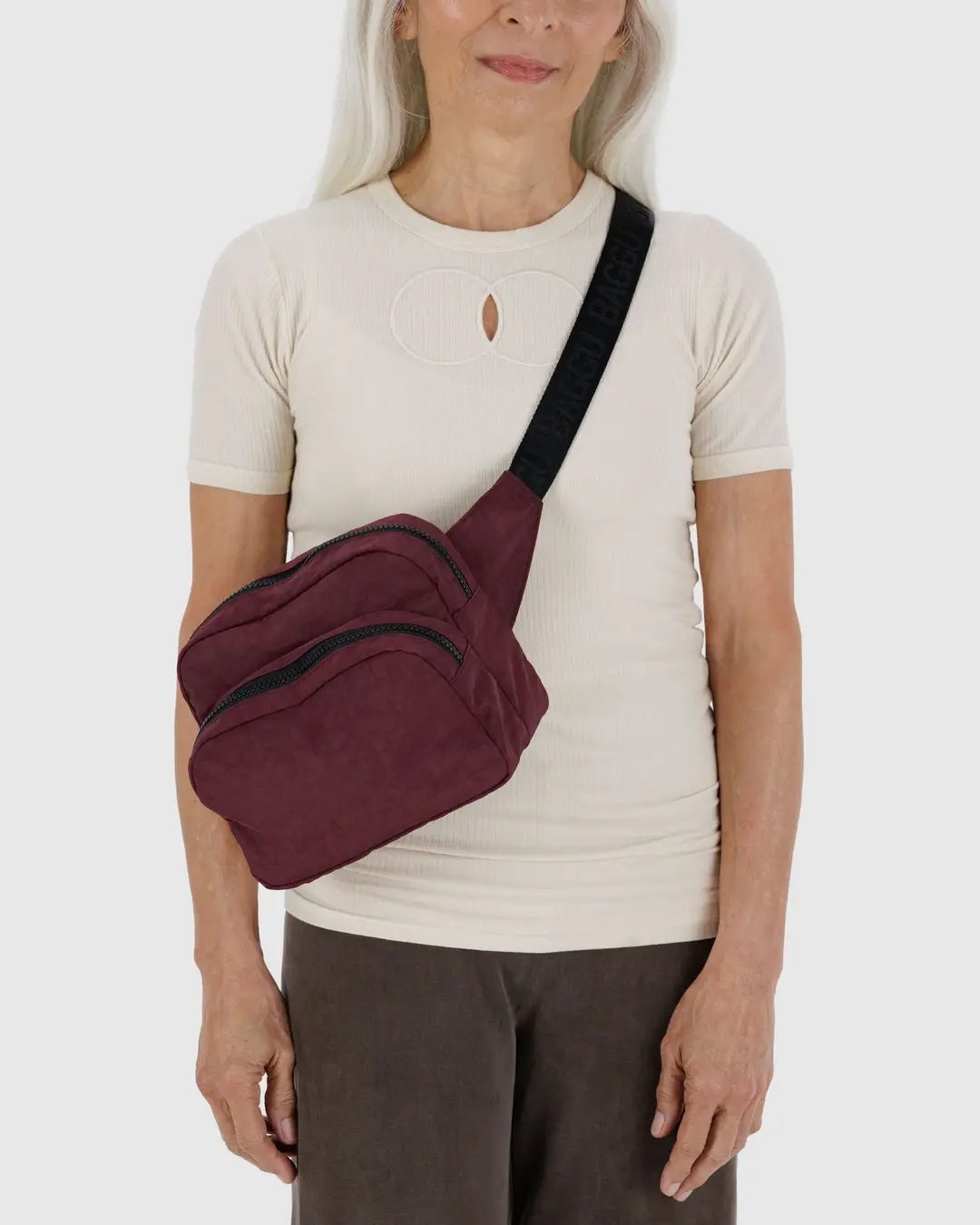 Person wearing a mahogany Baggu crossbody fanny pack bag standing in front of a white background