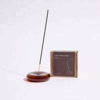 a round amber glass incense holder with a single incense stick next to its packaging on a white background