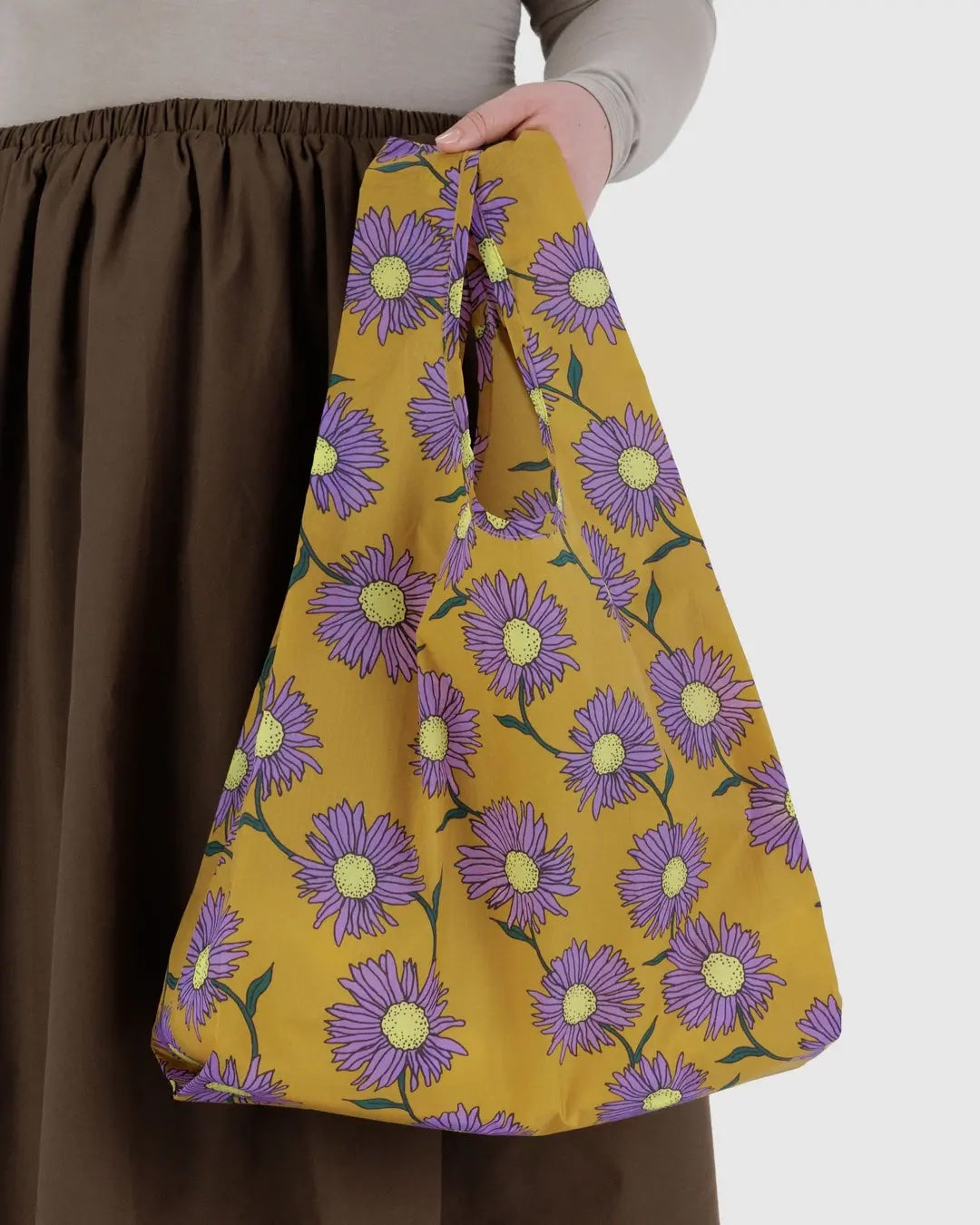 Baggu yellow reusable bag with purple floral pattern held by a person wearing brown pants.