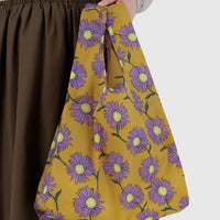 Baggu yellow reusable bag with purple floral pattern held by a person wearing brown pants.