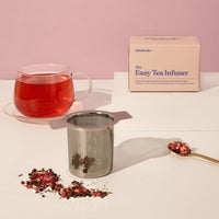 Tea cup with red liquid, tea infuser, and packaging on a light surface with pink background