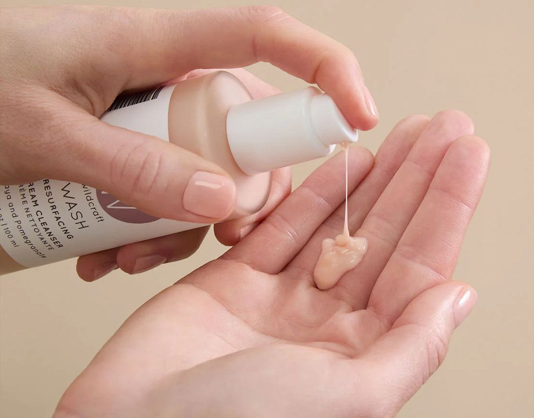 Hand holding a bottle of Wildcraft Cream Cleanser pumping product out onto their other hand against a beige background