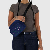 Person wearing a brown shirt and blue jeans wearing a baggu navy blue fanny pack bag with a pink embroidered floral pattern standing before a white background