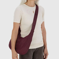 a person with long white hair wearing a white top and brown pants is shown wearing a medium Baggu crescent bag in mahogany red crossbody