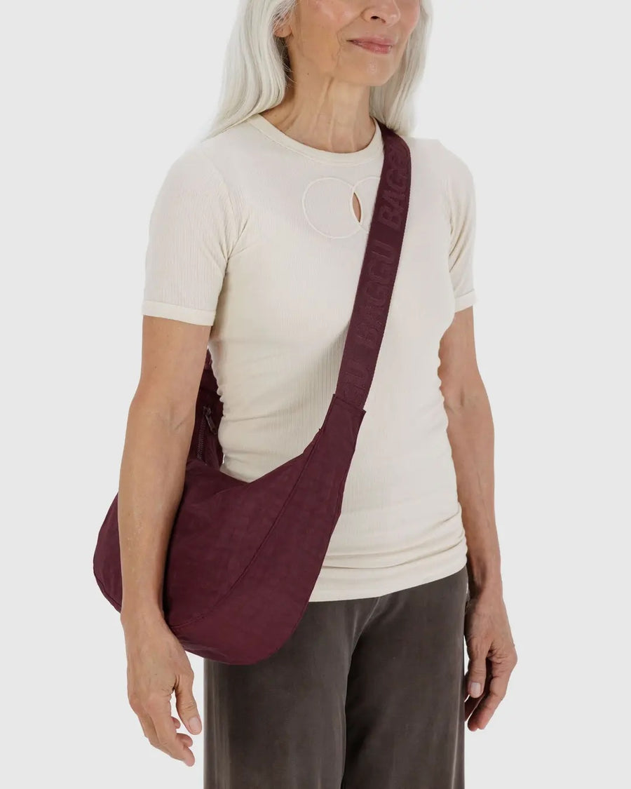 a person with long white hair wearing a white top and brown pants is shown wearing a medium Baggu crescent bag in mahogany red crossbody