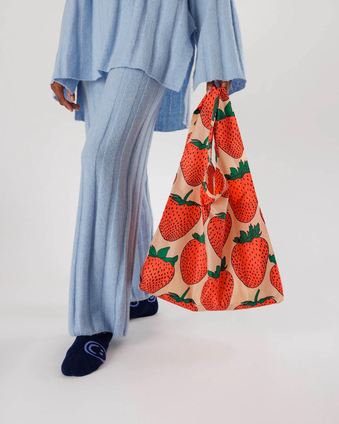 a person wearing a matching pale blue knitted sweater and pant set is shown from elbows down carrying a strawberry printed baggu reusable bag