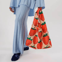 a person wearing a matching pale blue knitted sweater and pant set is shown from elbows down carrying a strawberry printed baggu reusable bag