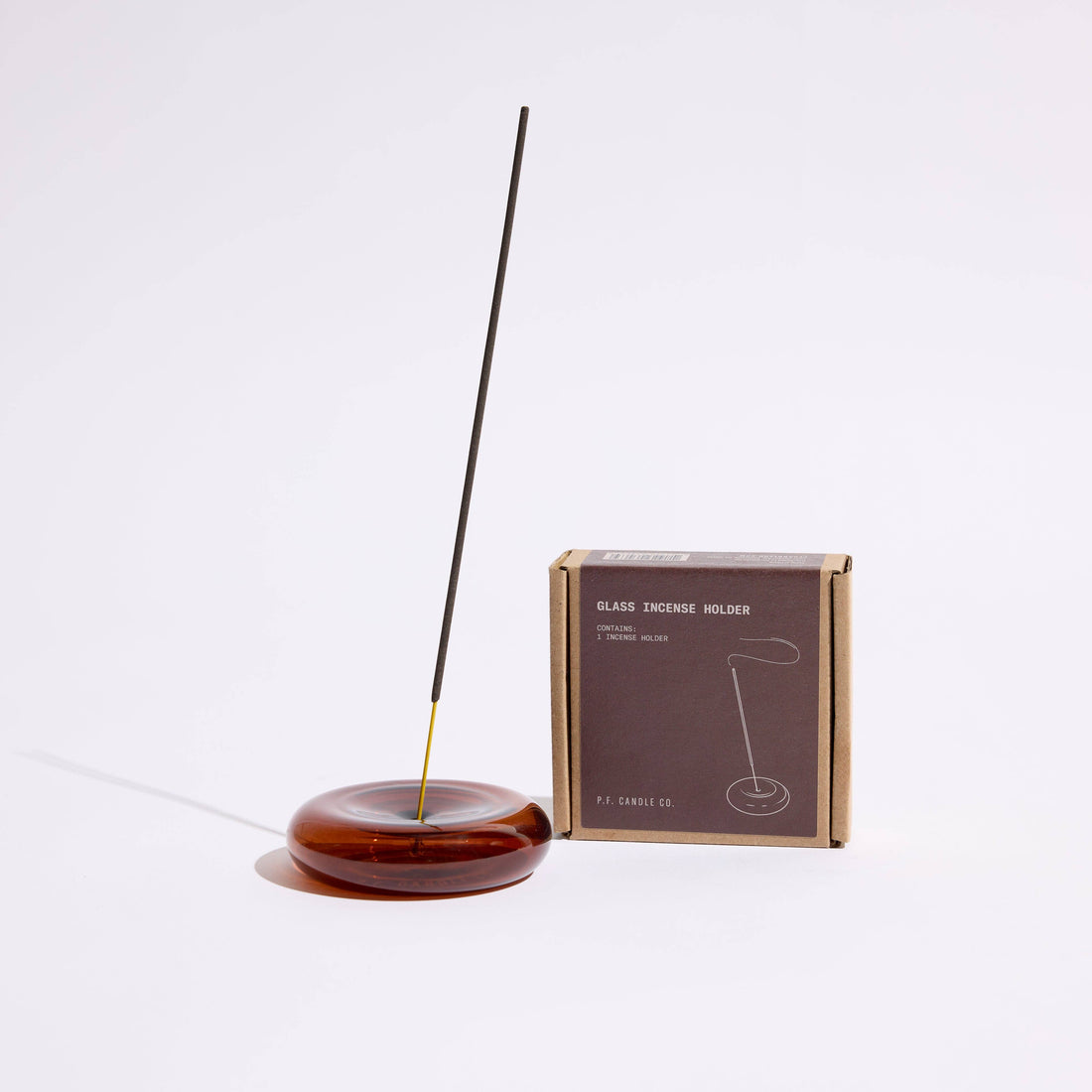 a round amber glass incense holder with a single incense stick next to its packaging on a white background