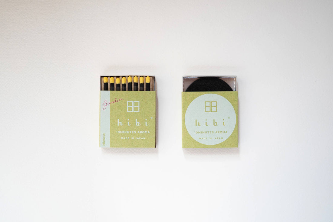 an open 8-pack of hibi mimosa incense matches in a green box on a white background.  The matches are black with canary yellow tips