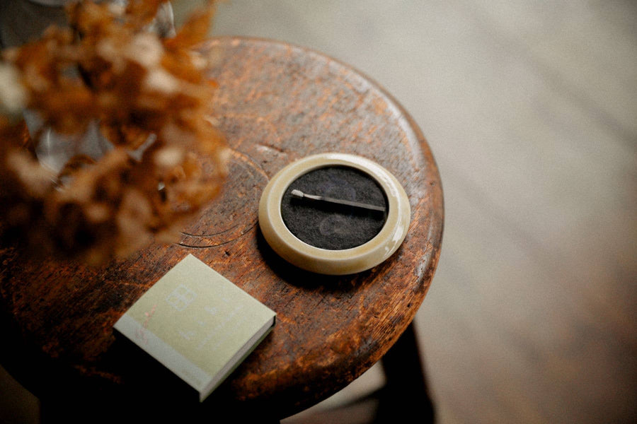 hibi yellow incense tray holding a burning incense match placed on a wooden stool with dried flowers and a pack of incense matches with a blurred floor background