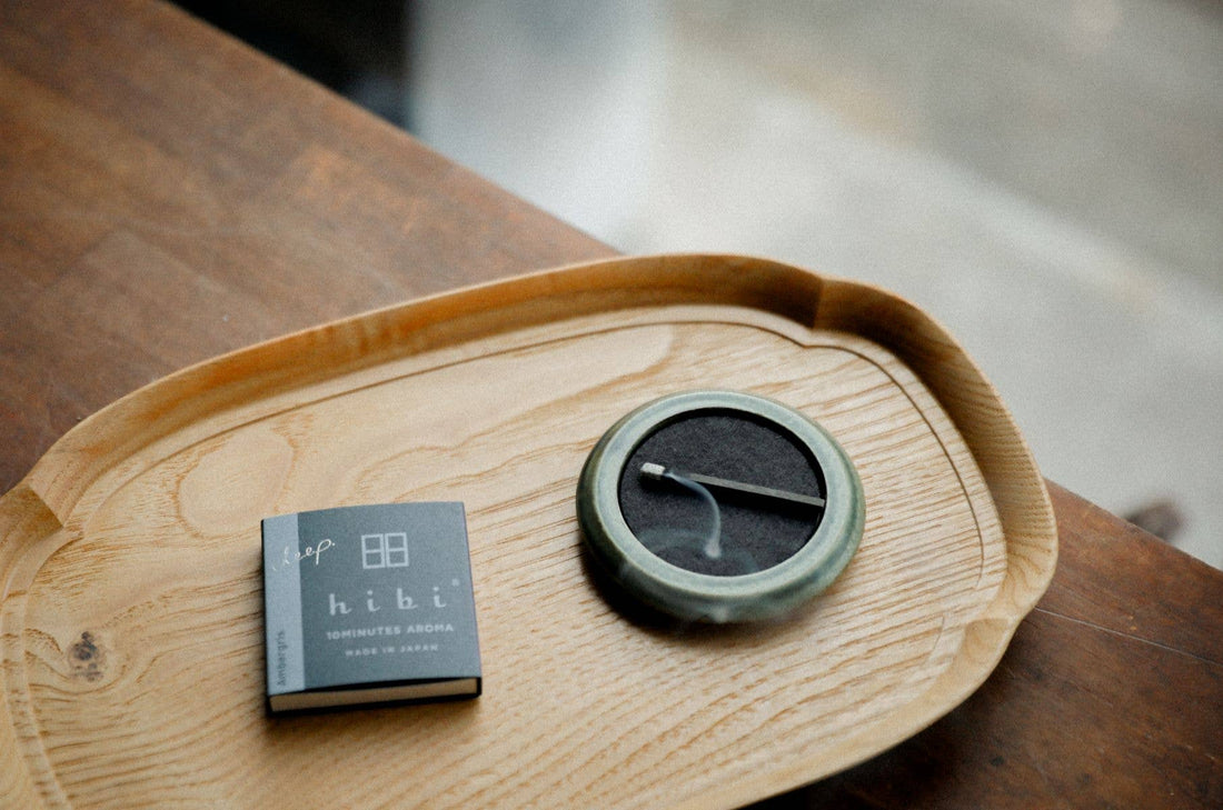 a small round green porcelain tray holding a burning hibi incense match along with a pack of the incense matches laying on a wooden tray on a table with blurred background 