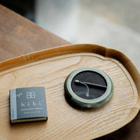 a small round green porcelain tray holding a burning hibi incense match along with a pack of the incense matches laying on a wooden tray on a table with blurred background 