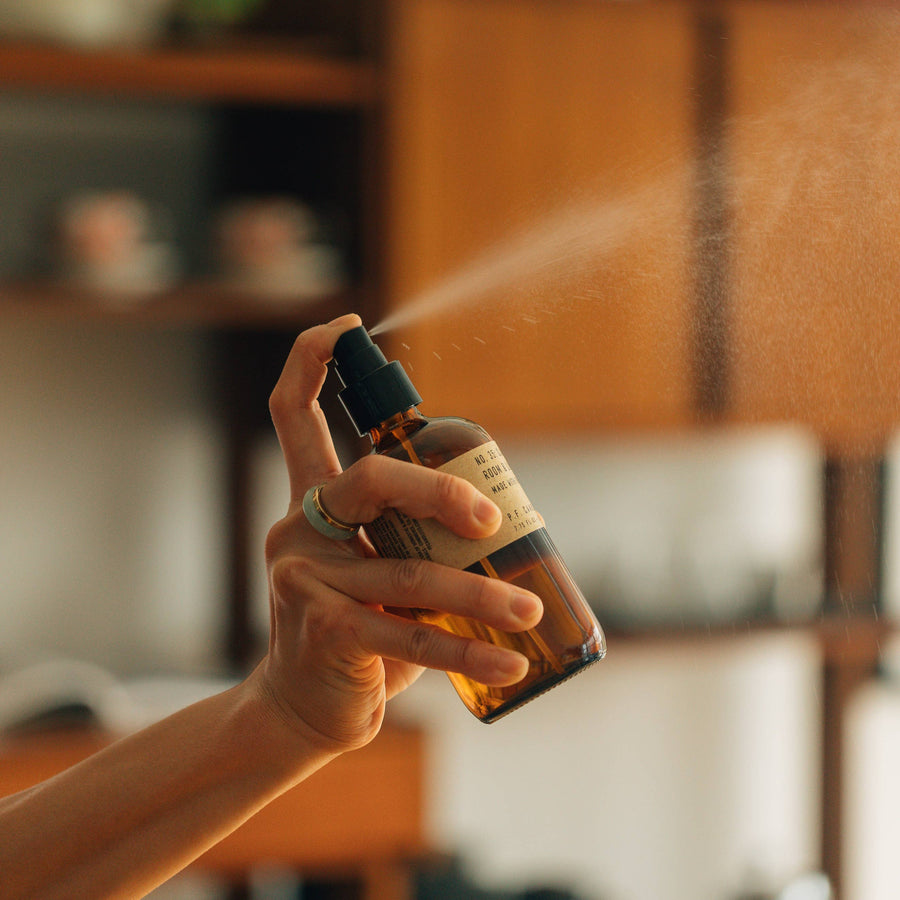 Hand holding an amber glass spray bottle spraying a liquid P.F. Candle Co Ojai Lavender Linen & Room Spray. There are warm wood cupboards and shelves blurred in the background.