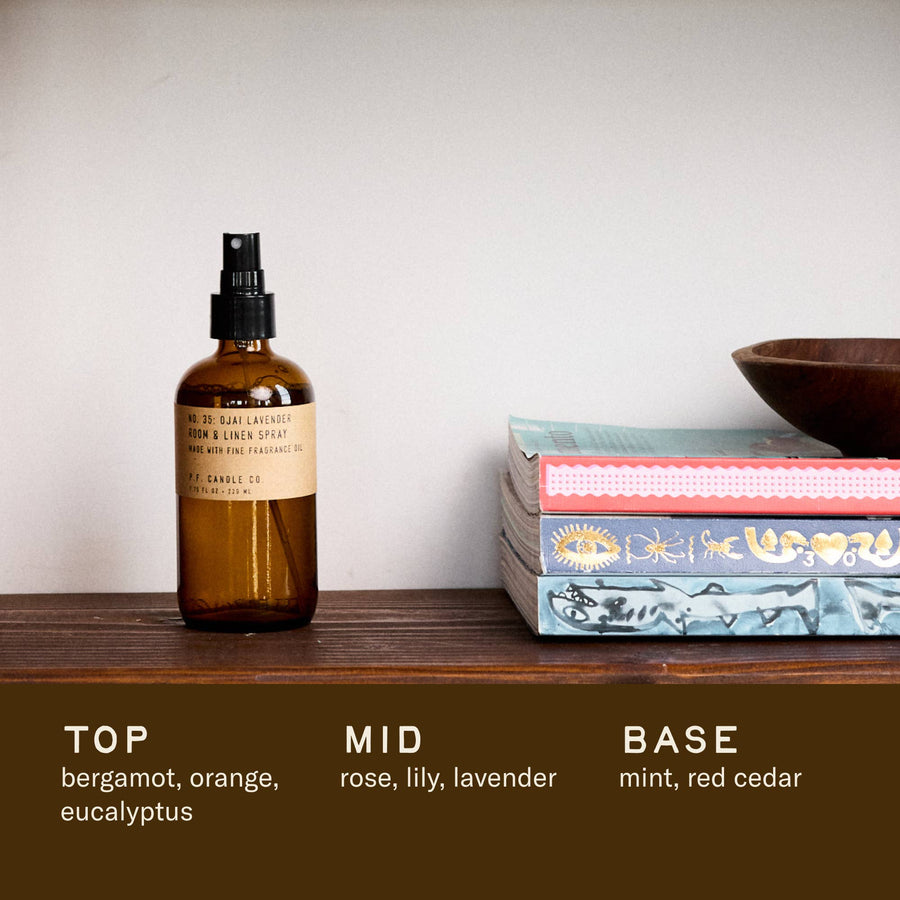 P.F. Candle Co Ojai Lavender spray in an amber glass spray bottle with a natural kraft label on a wooden surface with books and a bowl in the background.