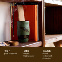 Pinon incense burning in a holder on a wooden shelf housing books and a green ceramic vessel.