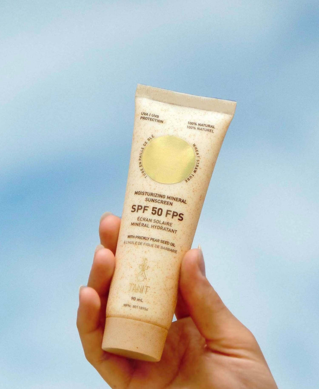 Hand holding a tube of Tannit SPF 50 sunscreen cream against a blue sky background