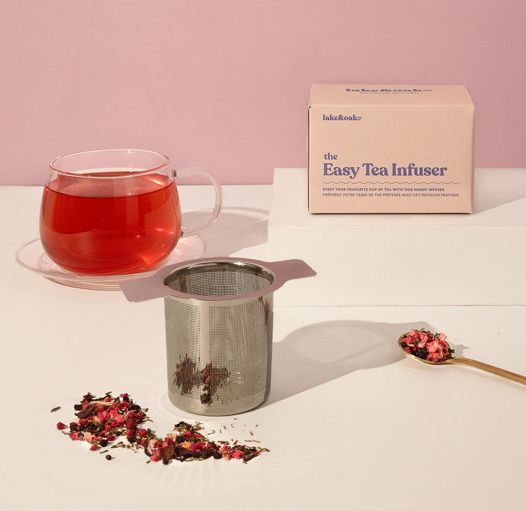Tea cup with red liquid, tea infuser, and packaging on a light surface with pink background