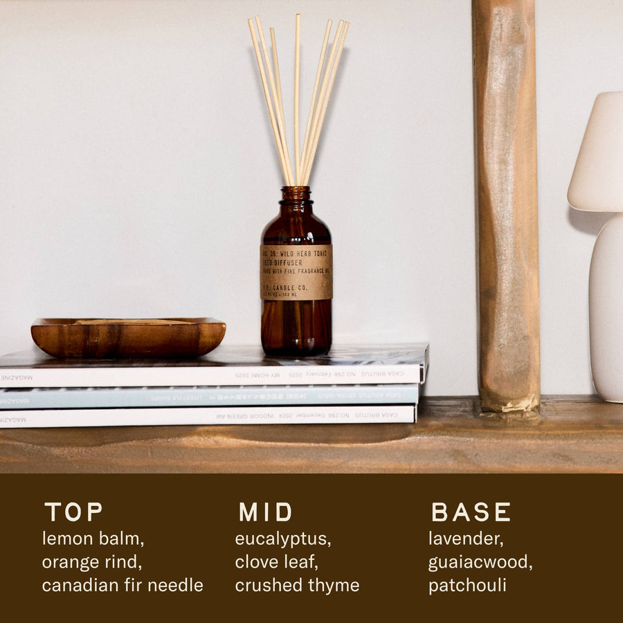 A P.F. Candle Co Wild Herb Tonic reed diffuser with wooden sticks sitting on a shelf with magazines and a small white lamp. The top, mid and base notes are written on the bottom of the image as follows. Top: lemon balm, orange rind, canadian fir. Mid: eucalyptus, clove leaf, thyme. Base: Lavender, guiacwood, patchouli