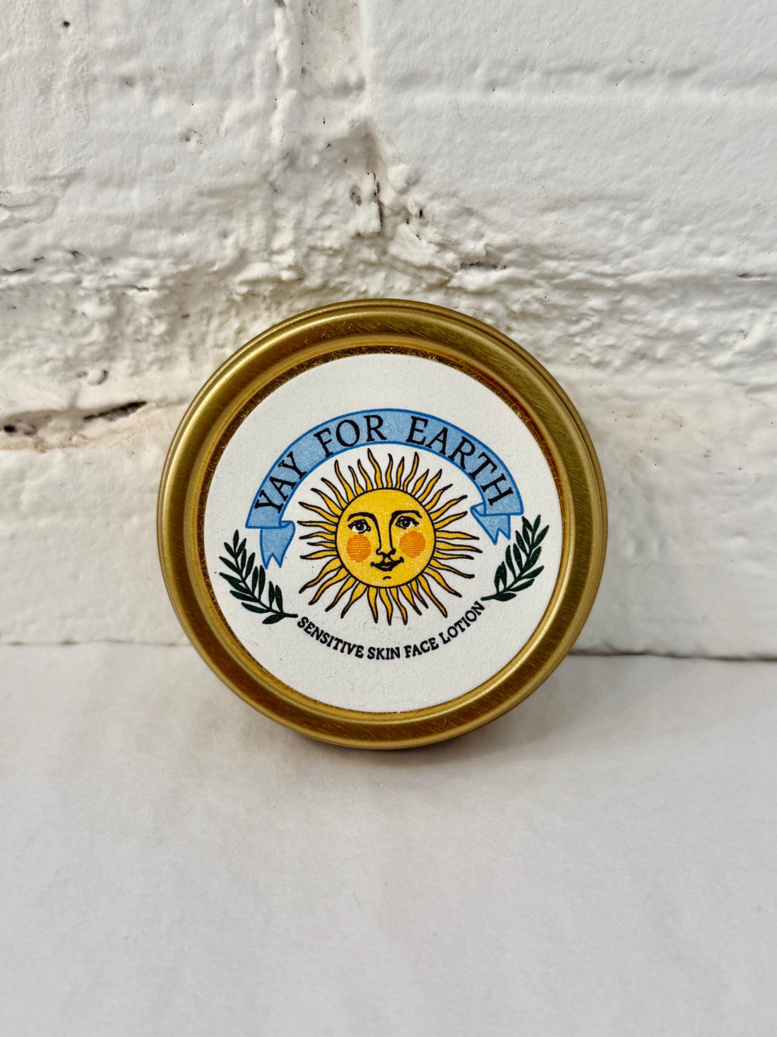 A round tin of 'Yay For Earth' sensitive skin face lotion with a gold-colored metal rim and a white label featuring a yellow sun and blue banner, set against a white painted brick wall.