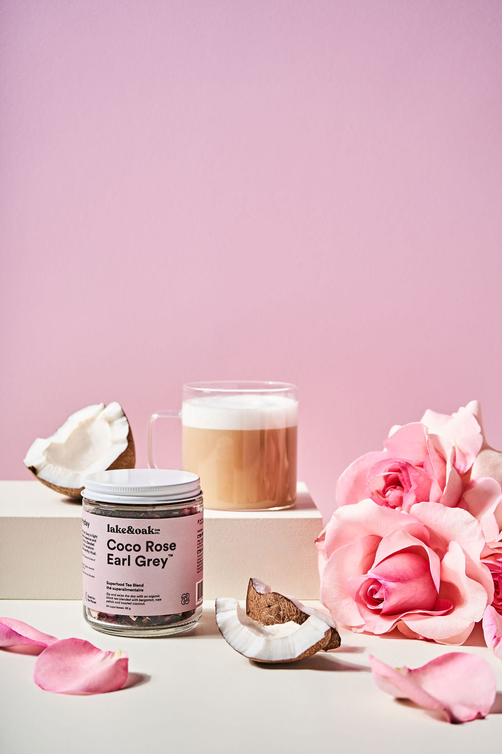a jar of Lake & Oak Coco Rose Earl Grey tea surrounded by lush pink roses and petals with a cup of tea with frothed milk in a clear glass mug and chunks of fresh coconut against a pink background. 