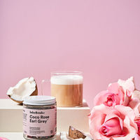 a jar of Lake & Oak Coco Rose Earl Grey tea surrounded by lush pink roses and petals with a cup of tea with frothed milk in a clear glass mug and chunks of fresh coconut against a pink background. 