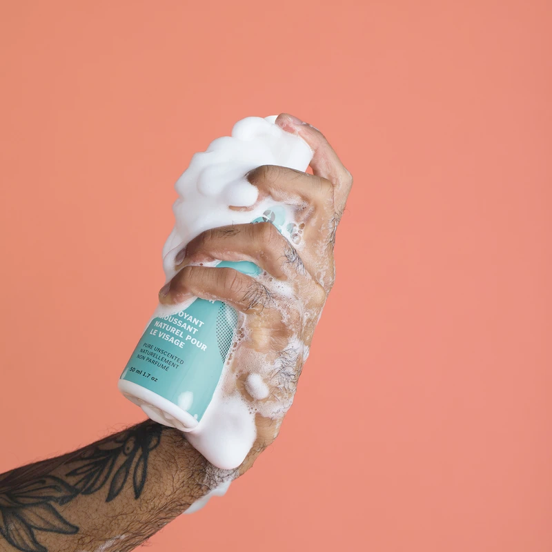 a hand is holding a bottle of Consonant Natural Foaming Face Wash covered in rich foamy lather
